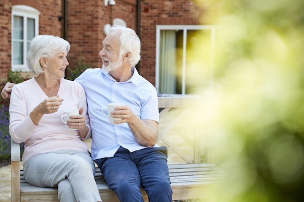 Senior Couple Sitting On Bench With Hot Drink In Assisted Living Facility